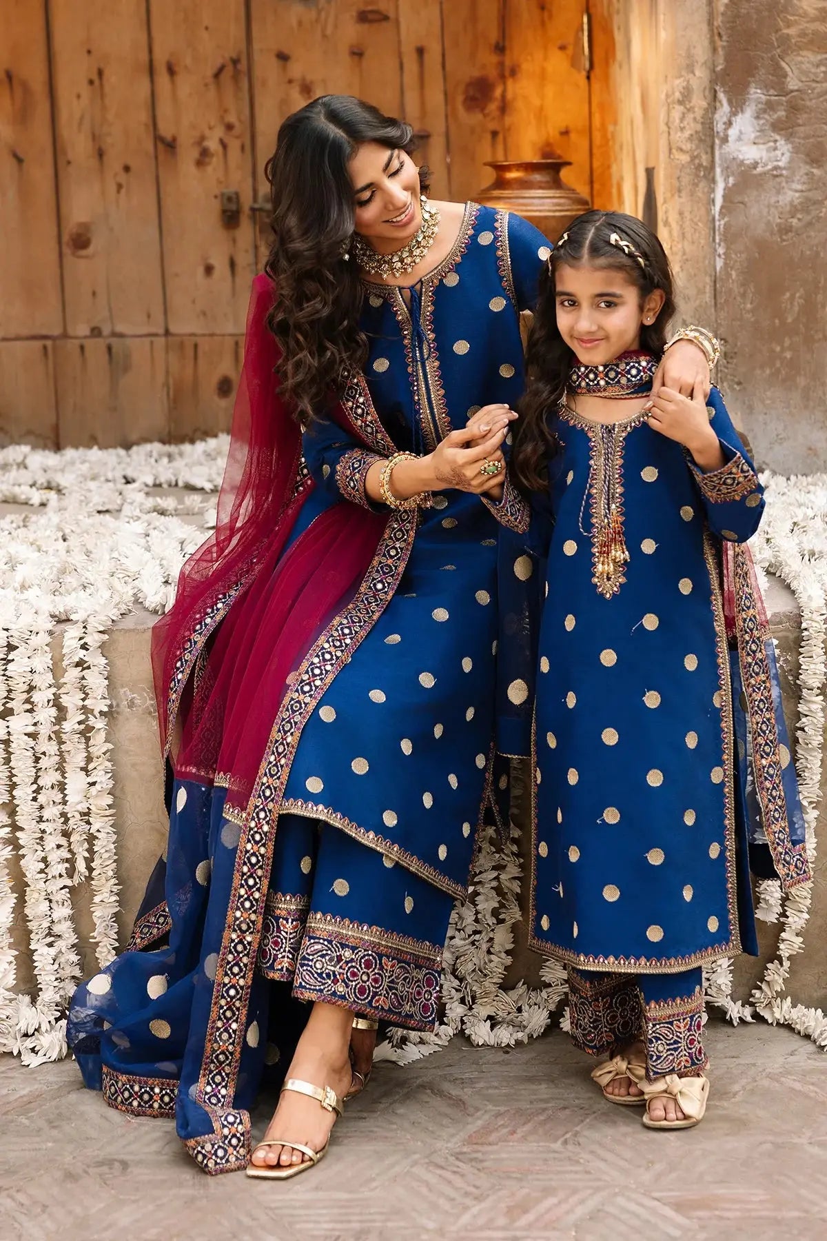Woman and young girl in matching blue traditional outfits with white polka dots, standing against a wooden wall.