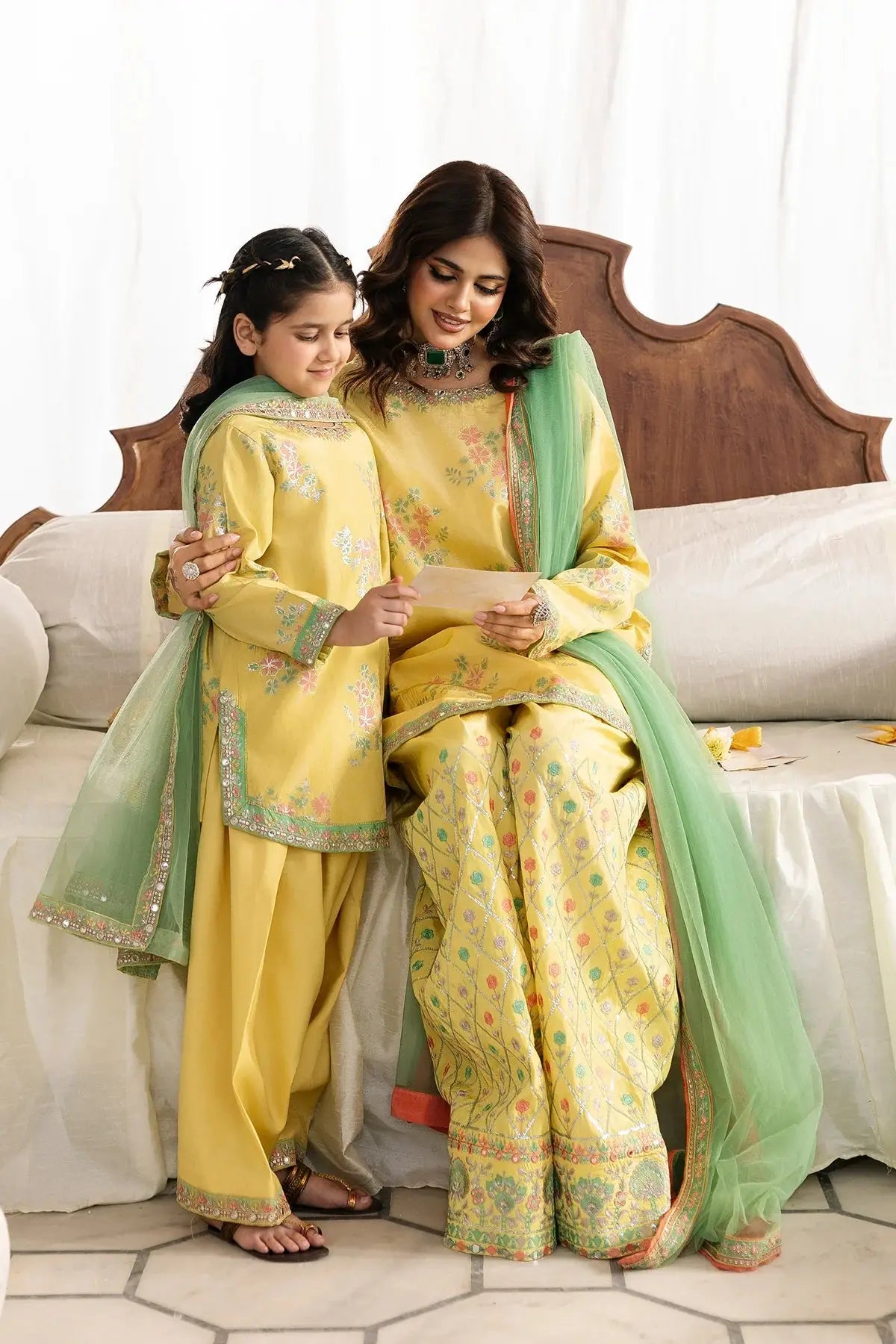 Two women in matching yellow and green traditional outfits sitting on a couch.
