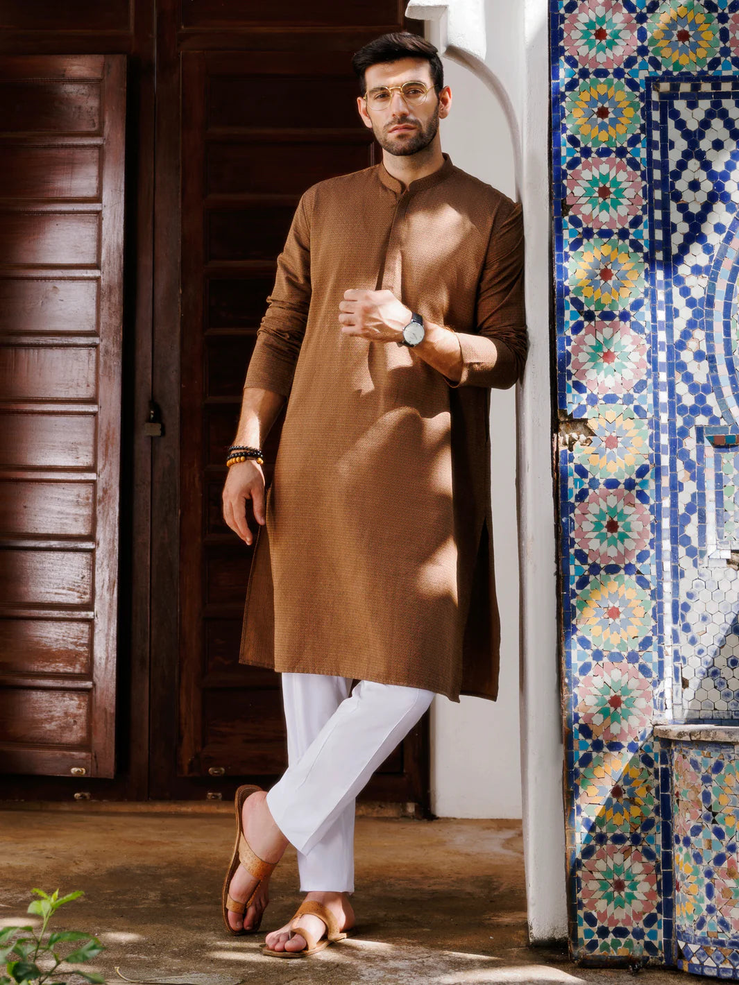 Men's brown kurta with white pants, standing against wooden and mosaic background, casual style