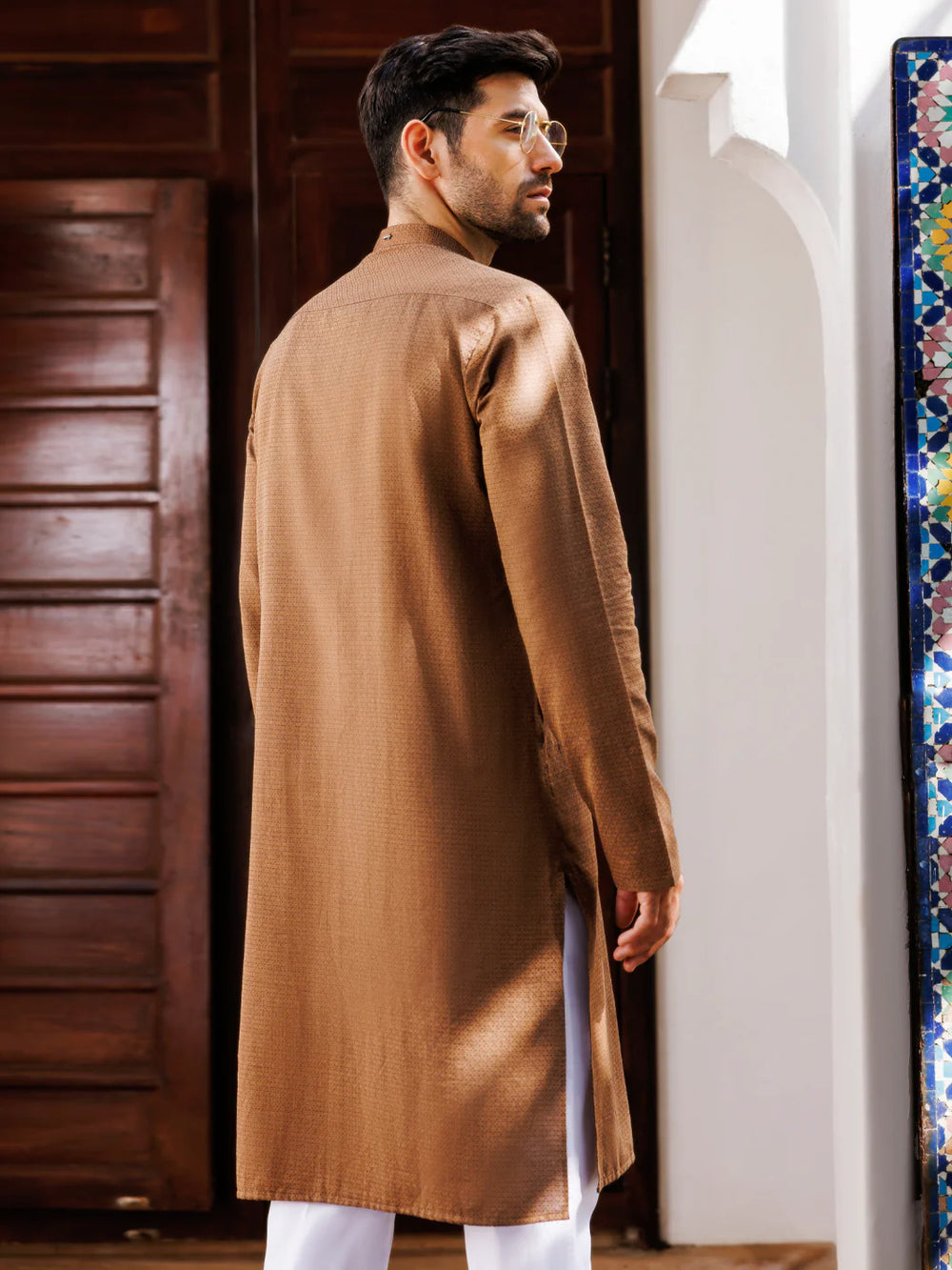 Back view of man in brown kurta with white pants, standing near wooden door and mosaic wall, stylish look