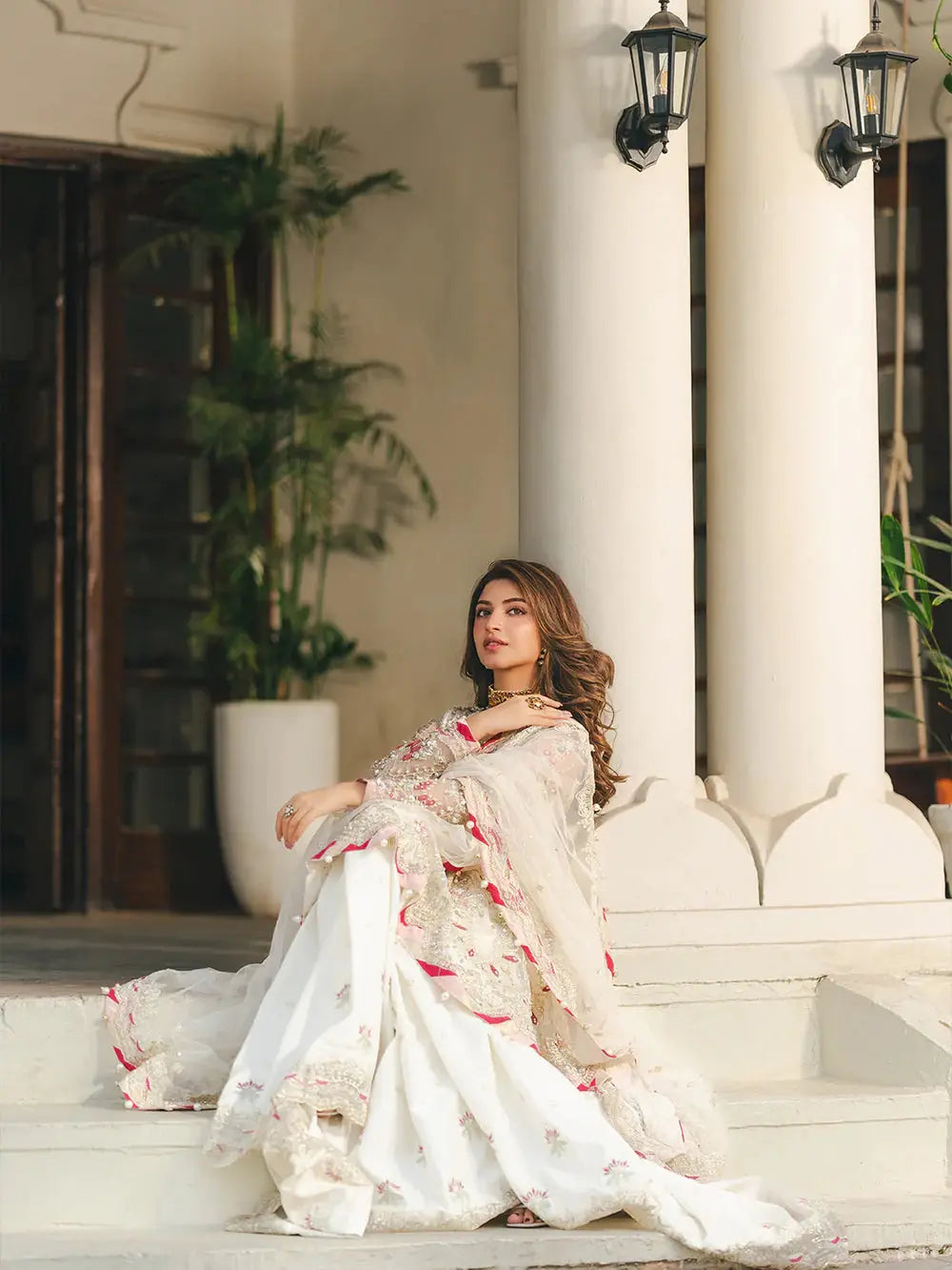Model sitting on white steps in traditional embroidered outfit with lush indoor plants and architectural columns, showcasing elegant bridal attire