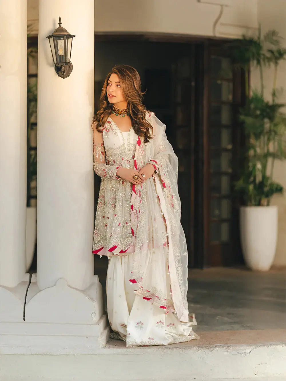 Beautiful woman in white embroidered traditional dress with intricate embellishments, wearing jewelry, standing near a column for wedding or festive occasion