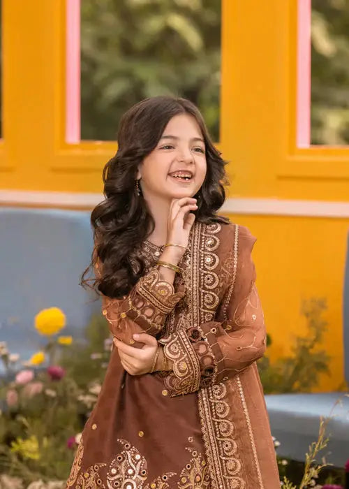 Smiling girl in brown embroidered dress with dupatta, standing in garden with yellow wall background