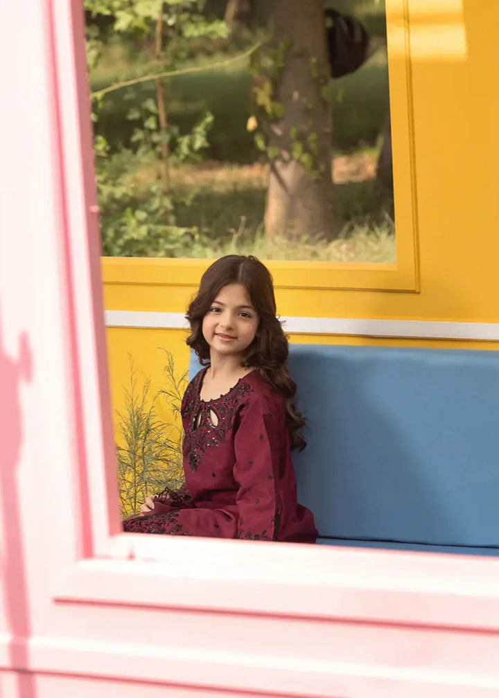 Young girl in maroon embroidered dress sitting on blue bench near yellow window
