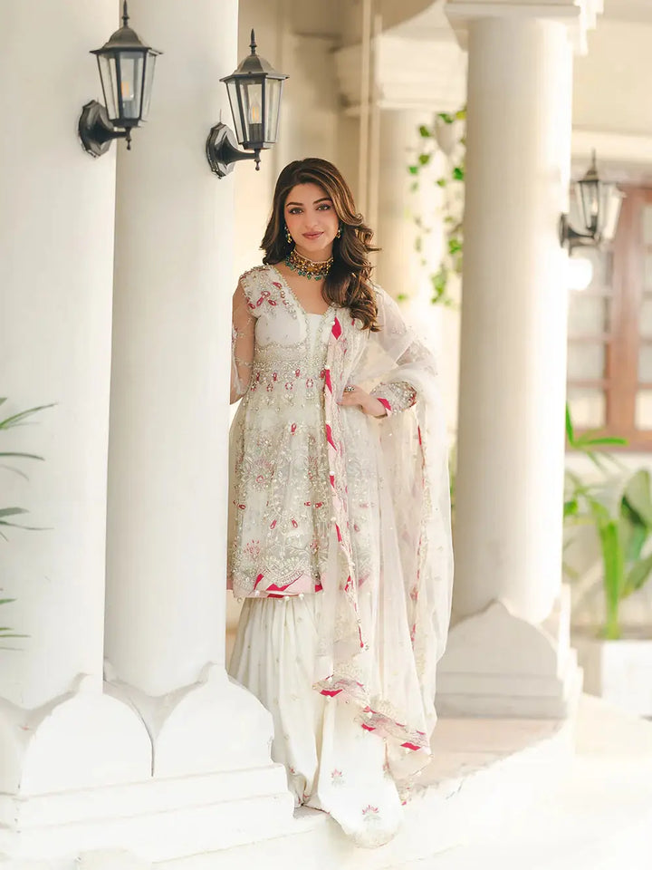 Stylish woman in white embroidered dress with detailed embellishments, posing near a column with a smile, showcasing wedding or festive outfit