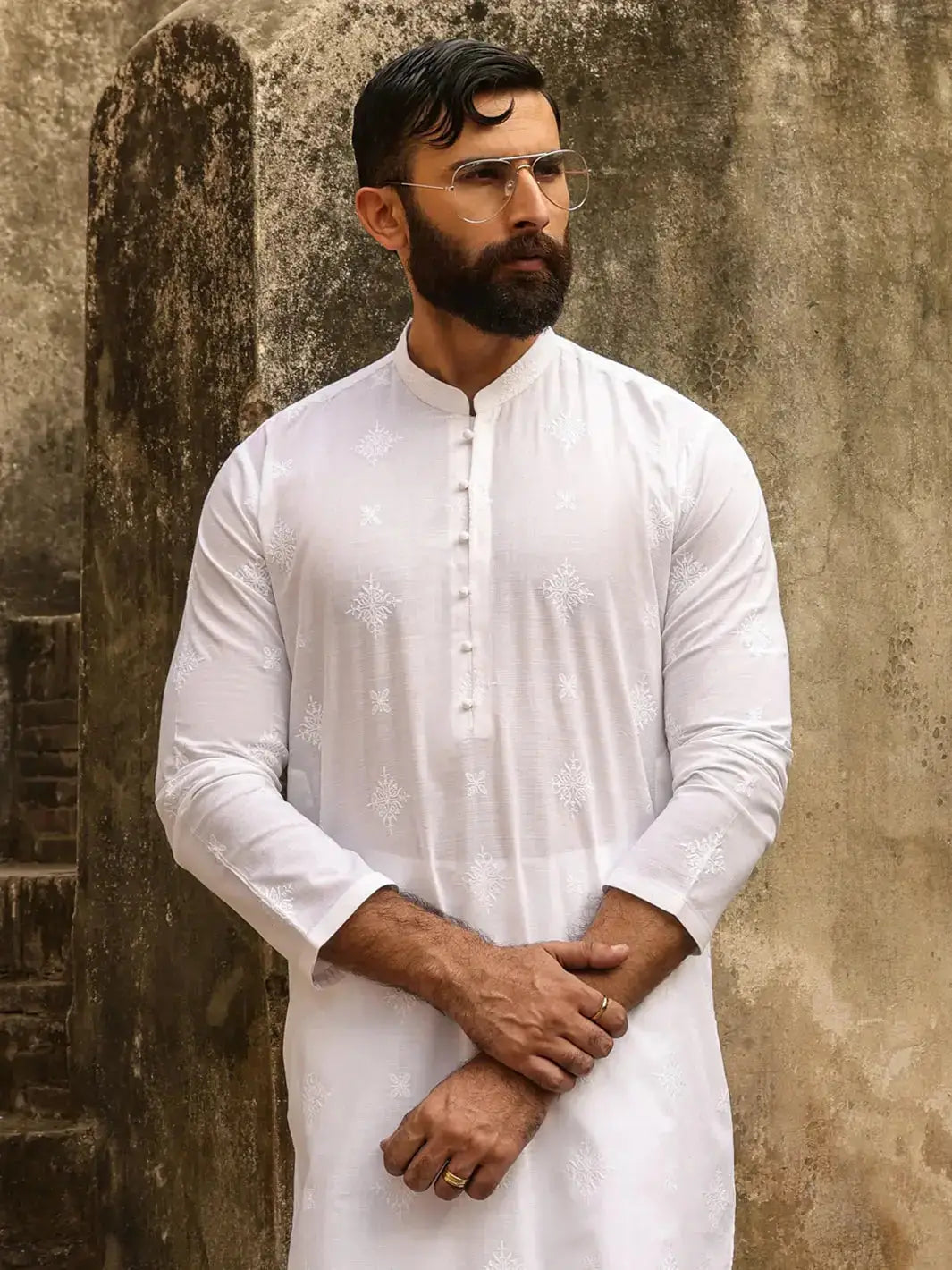 Man in white embroidered kurta with fabric buttons, posing against textured wall