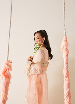 Model in MARYUM AND MARIA ELYNA chiffon outfit holding flowers, showcasing soft peach tones and traditional Pakistani design.