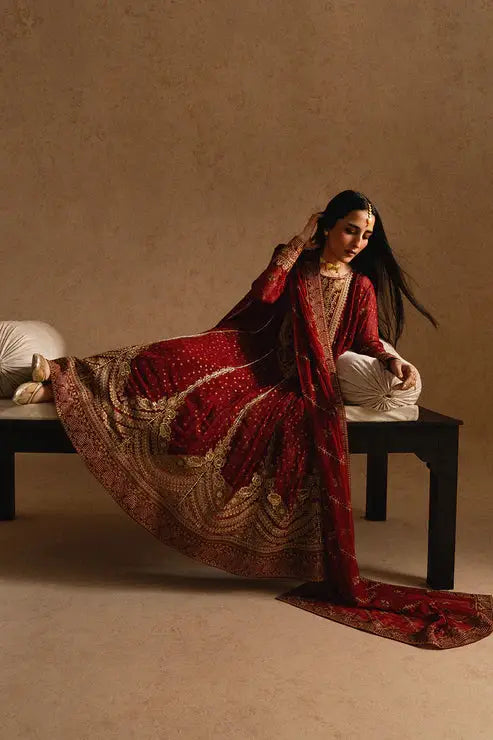 Model lounging in MARYUM AND MARIA MELISA embroidered chiffon dress, showcasing full outfit with ornate gold patterns on maroon fabric.