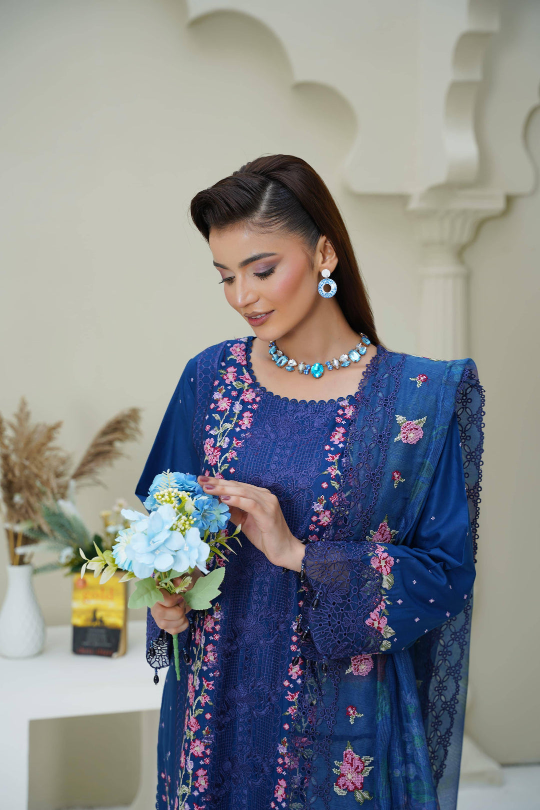 Model in RANGREZA SAJNI RS-3518-1000 admiring flowers, wearing a blue embroidered Pakistani suit with matching jewelry for a festive look.