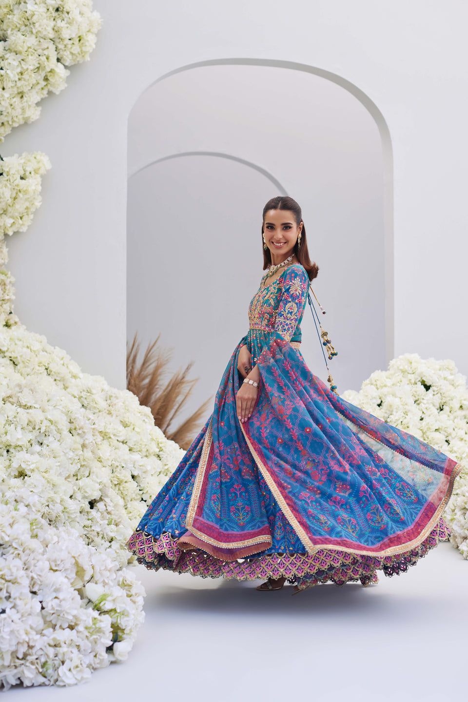 Mina Hasan shirt, bottom, and dupatta set in blue with intricate floral embroidery, worn in a twirling pose among white floral decor.