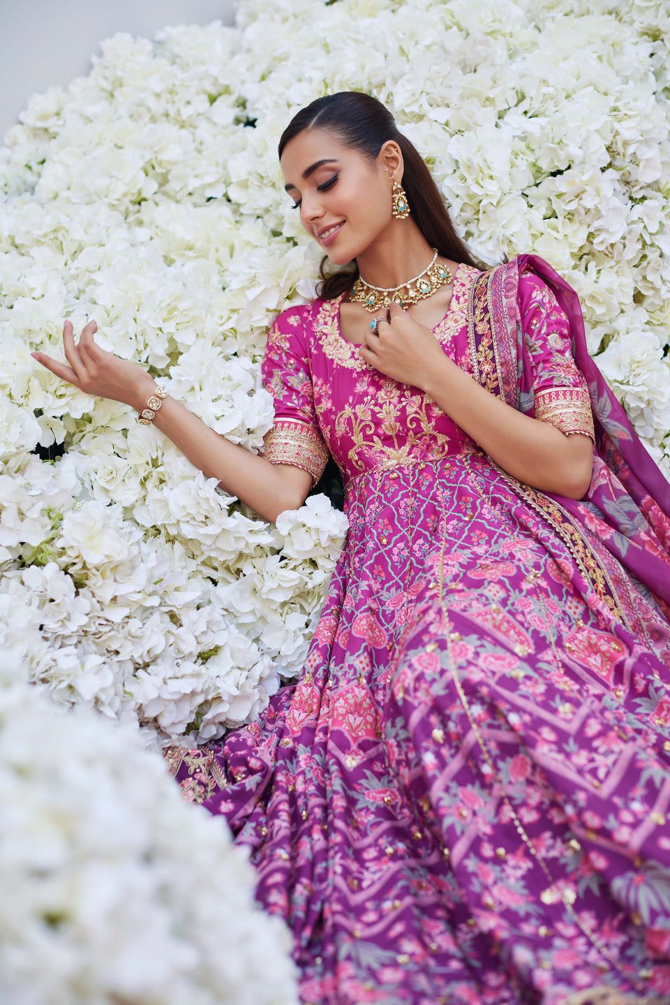 Mina Hasan pink ombre embroidered outfit with floral and geometric patterns, model reclining on white hydrangeas in a dreamy pose.