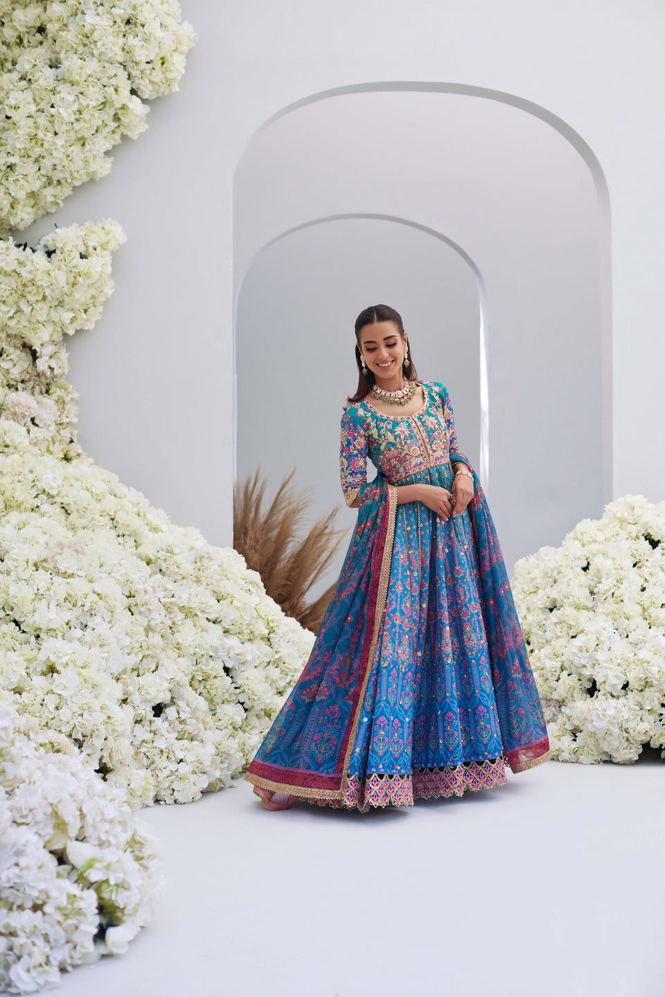 Full view of Mina Hasan blue embroidered outfit with pink accents, model posing gracefully among white floral arrangements.