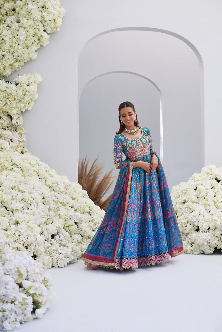 Full view of Mina Hasan blue embroidered outfit with pink accents, model posing gracefully among white floral arrangements.
