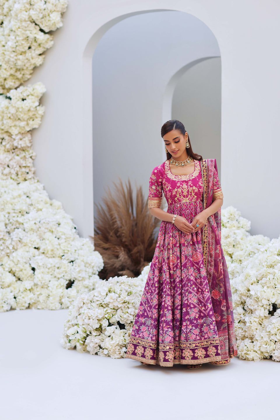 Mina Hasan pink ombre embroidered outfit with gold detailing, model posing gracefully in a white floral background.