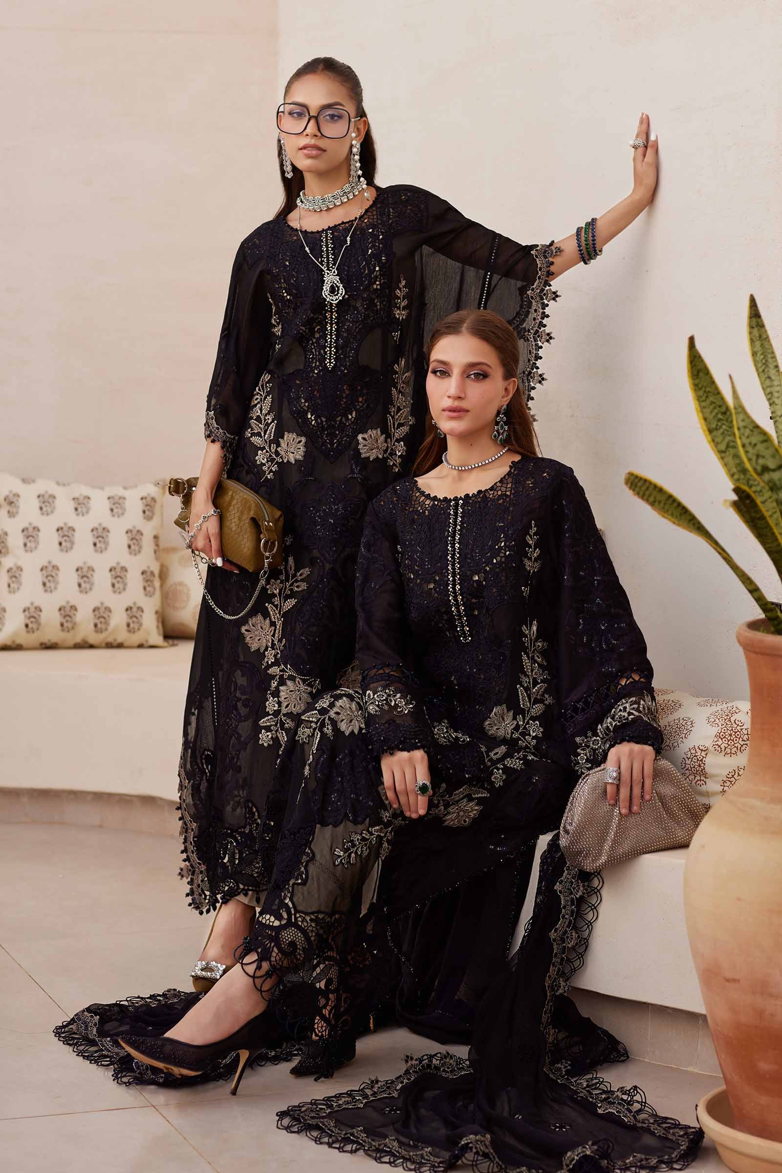 Two women in black embroidered dresses posing in a room with a plant and couch.