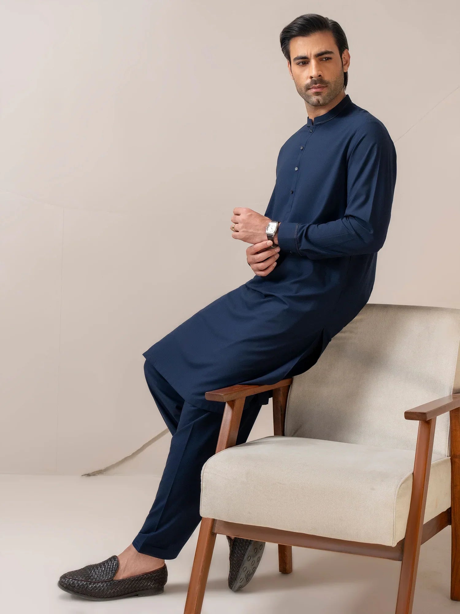 Man sitting on a chair wearing a navy blue kurta with white pants against a beige background