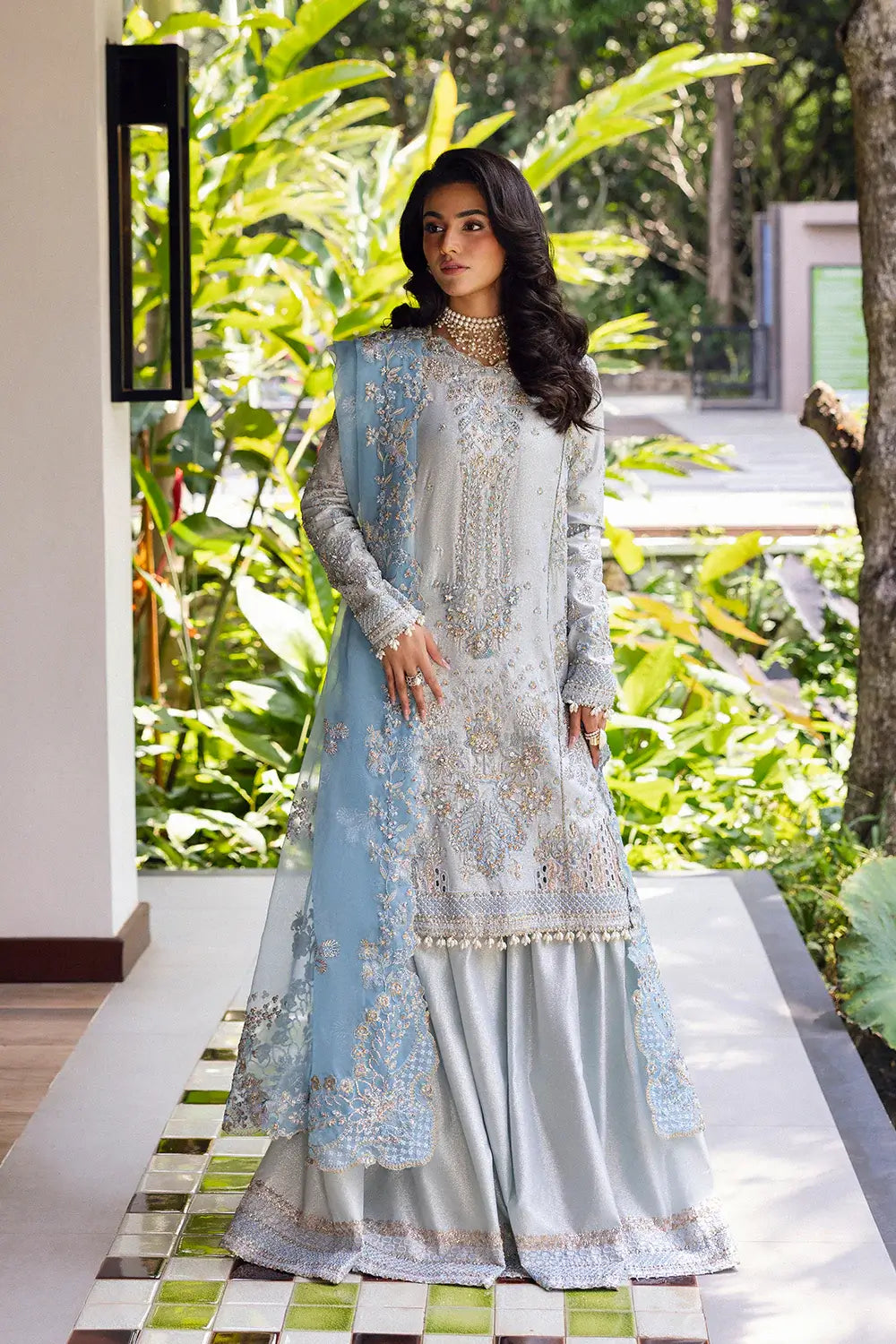 Woman in a light blue and white embroidered traditional outfit standing outdoors with greenery.