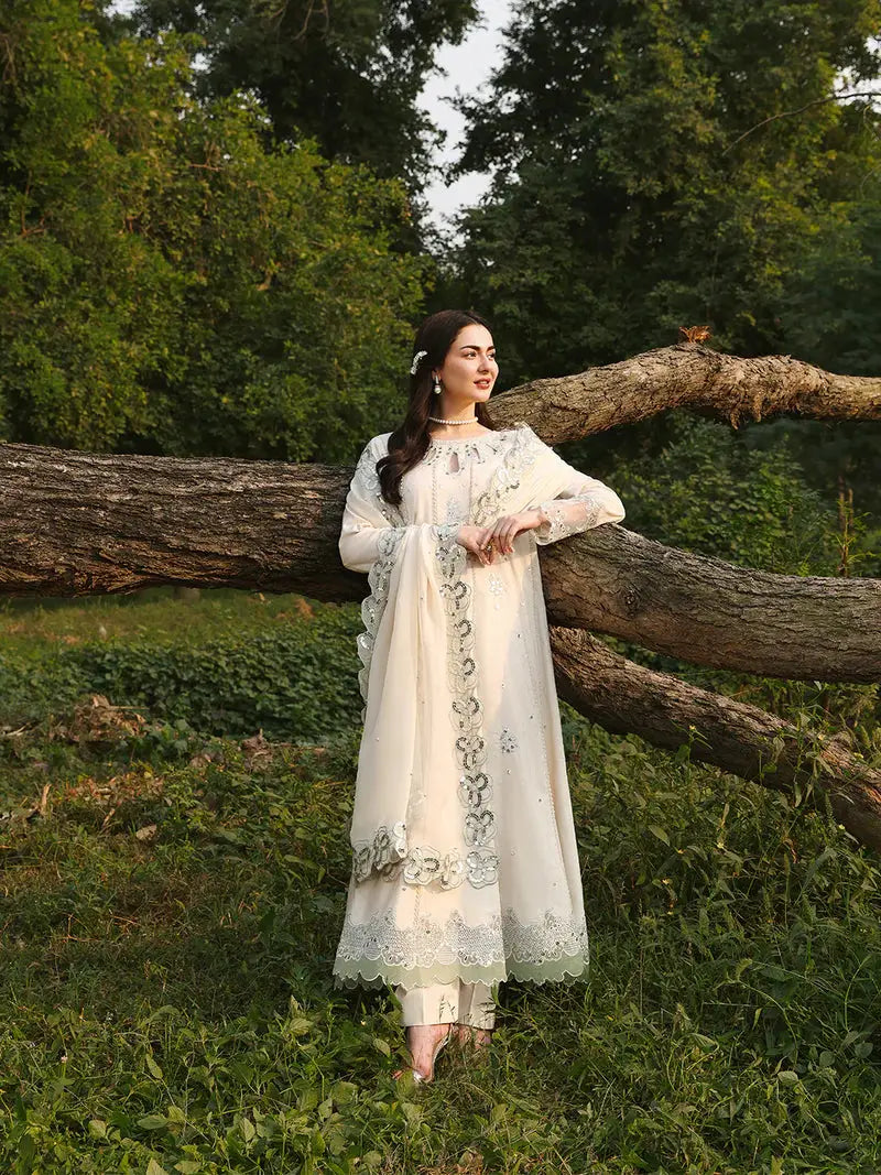 Woman in cream embroidered dress with dupatta, floral border, and lace, standing outdoors near a tree
