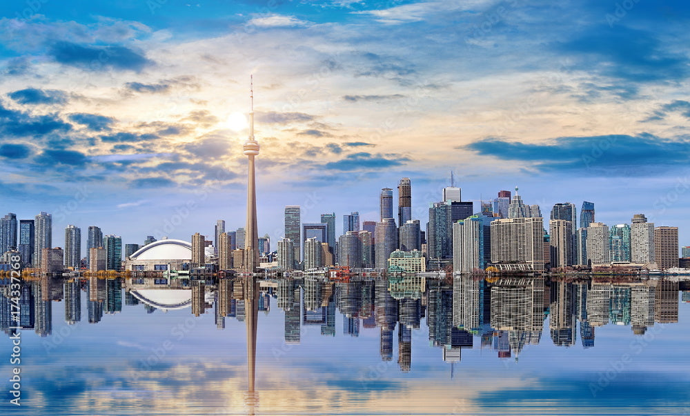 Toronto skyline at sunset with the CN Tower and surrounding skyscrapers reflected in the calm waters of Lake Ontario under a partly cloudy sky.