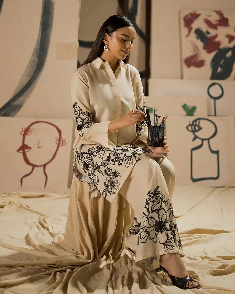 Model seated in EA-19 Pakistani outfit, showcasing detailed black floral prints on cream raw silk shirt and pants, ideal for Eid events.