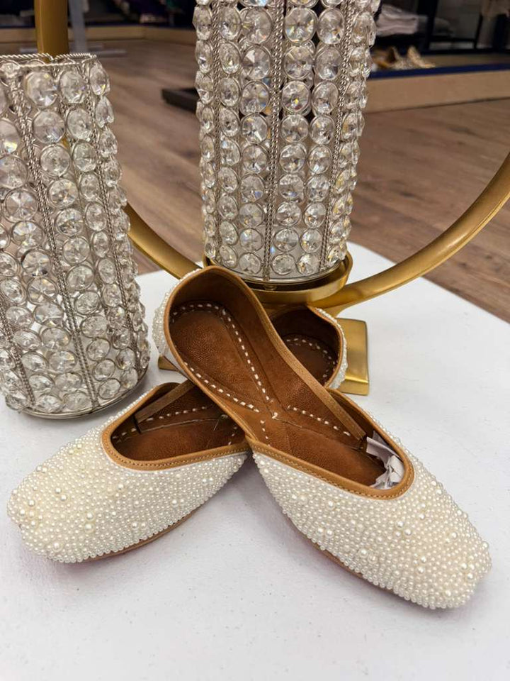 White Khussa Jutti for weddings with pearl embellishments, displayed in front of crystal candle holders. Traditional slip-on bridal flats.