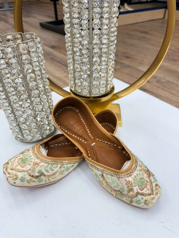 Mehrunnisa Gold Work Jutti with floral mirror embroidery, displayed on a white surface beside crystal decor. Shop Khussa Pakistan flats online.