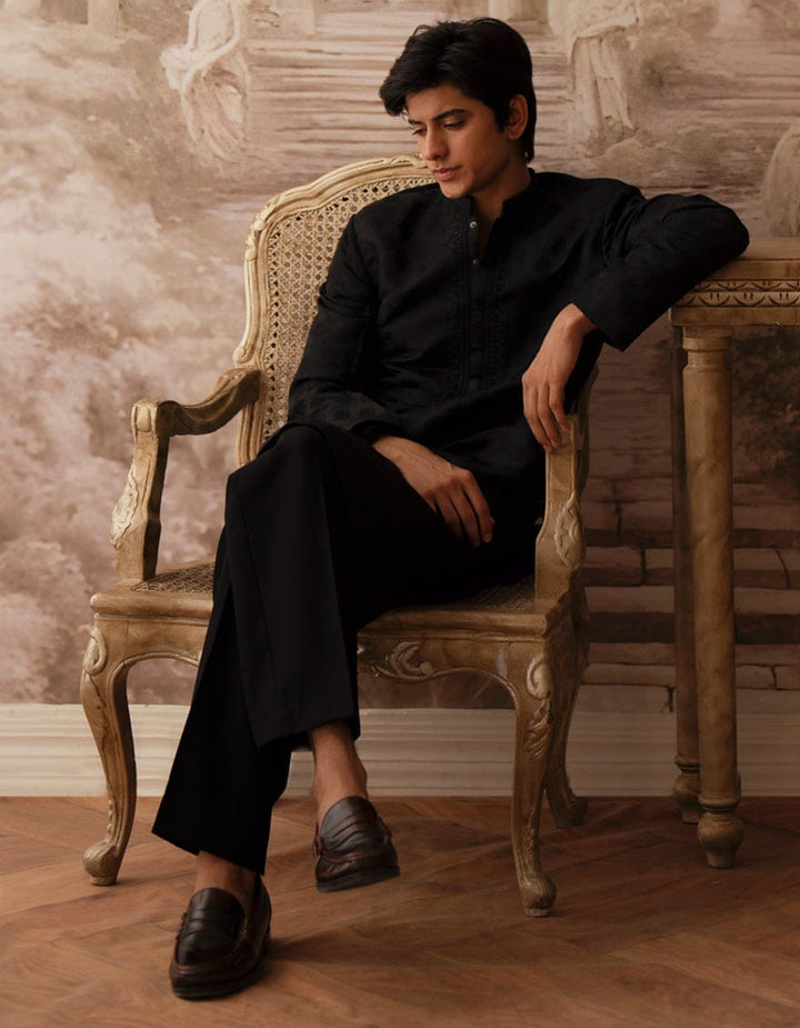 Model seated wearing J. BLACK BLENDED boys kurta in black, styled with black trousers and loafers for a refined traditional look.