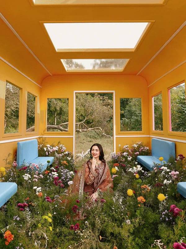 Woman in brown embroidered traditional dress sitting among flowers in a yellow room with large windows and greenery outside