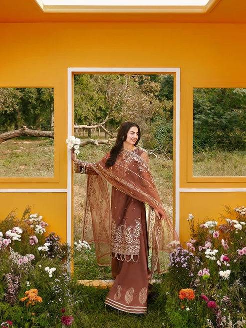 Woman in brown embroidered traditional dress posing with flowers in a yellow room with large windows and outdoor scenery