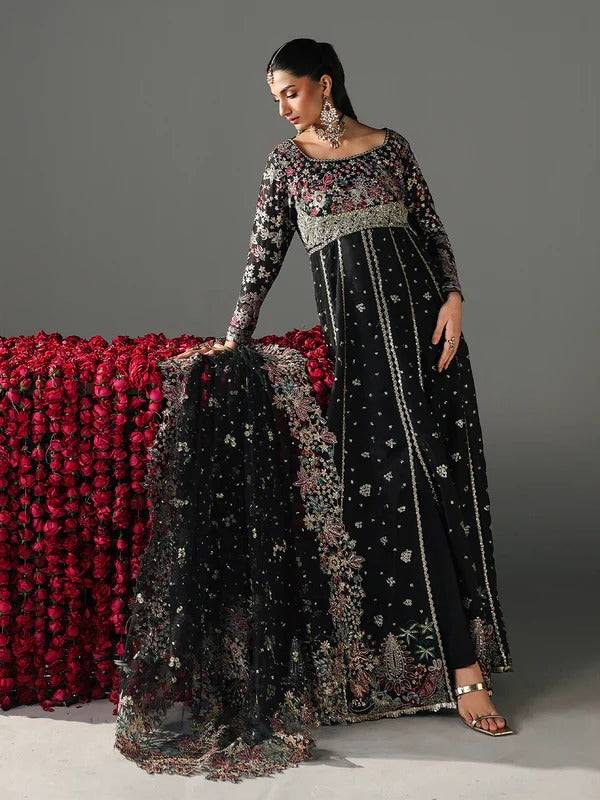 Model in black embroidered dress with floral and intricate embroidery, standing beside red floral backdrop