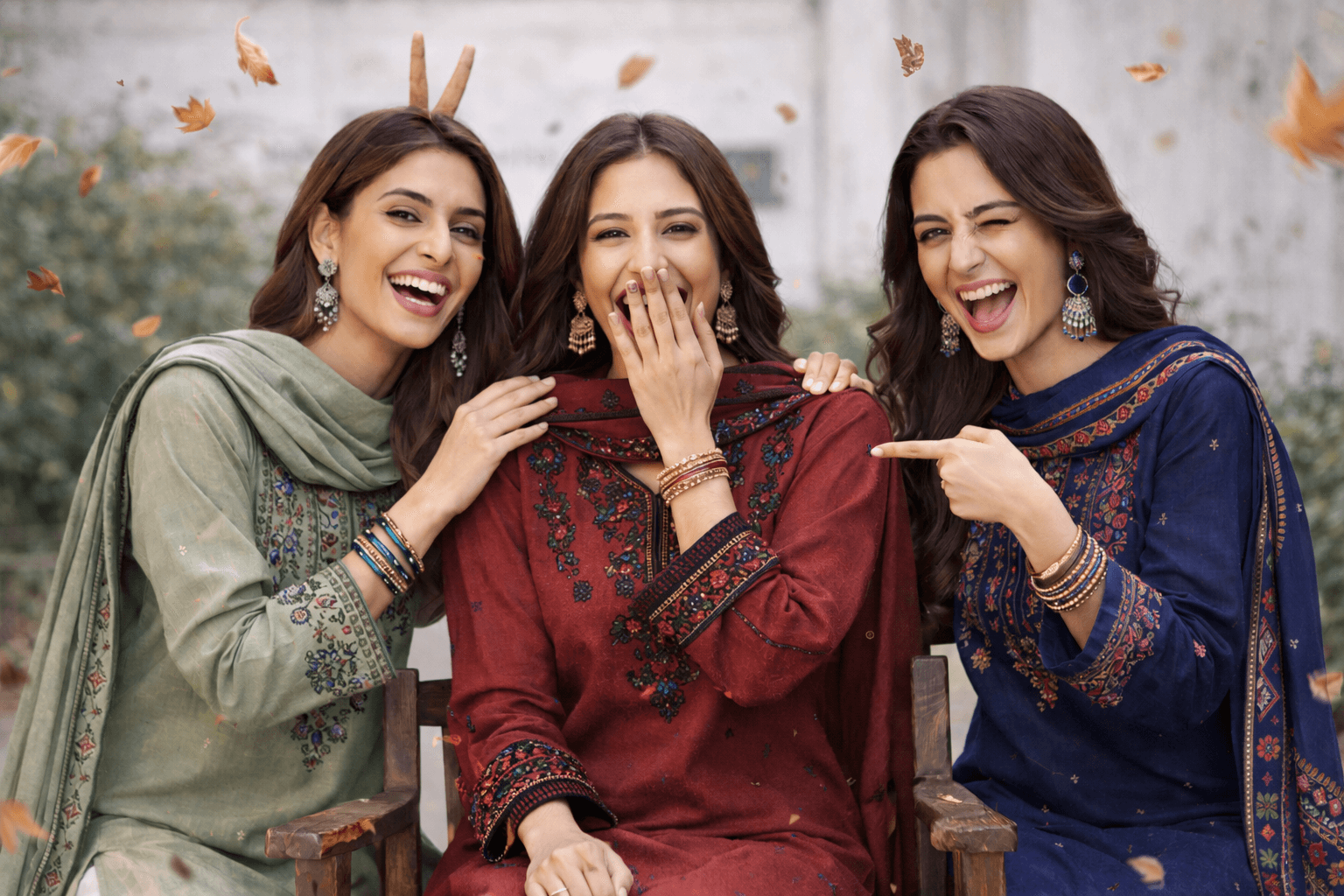 Three women in traditional attire laughing together outdoors with autumn leaves falling.